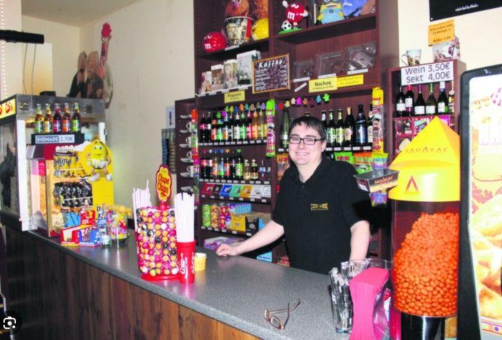 A smiling young man stands behind a busy counter filled with snacks and drinks.